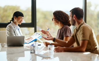A business advisor in a suit gestures while explaining documents to a seated couple during an indoor meeting, with papers and a laptop visible on the table.